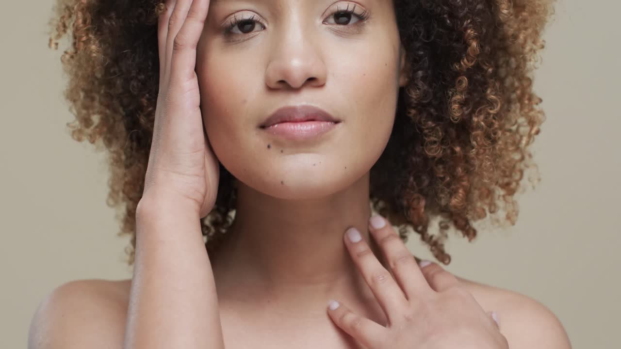 Mulher birracial com cabelo escuro tocando o rosto em fundo bege, em câmera lenta