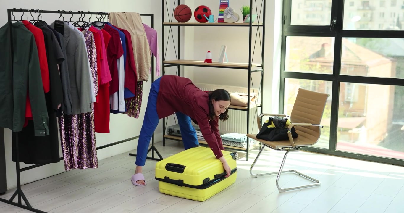 Woman in jacket bends over trying to close bright yellow suitcase