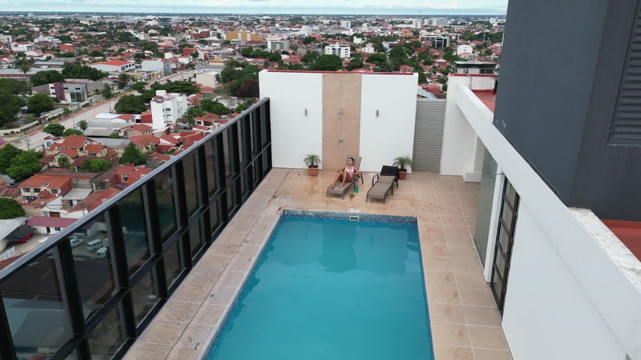 Aerial pulls back: Woman sitting by rooftop swimming pool on highrise