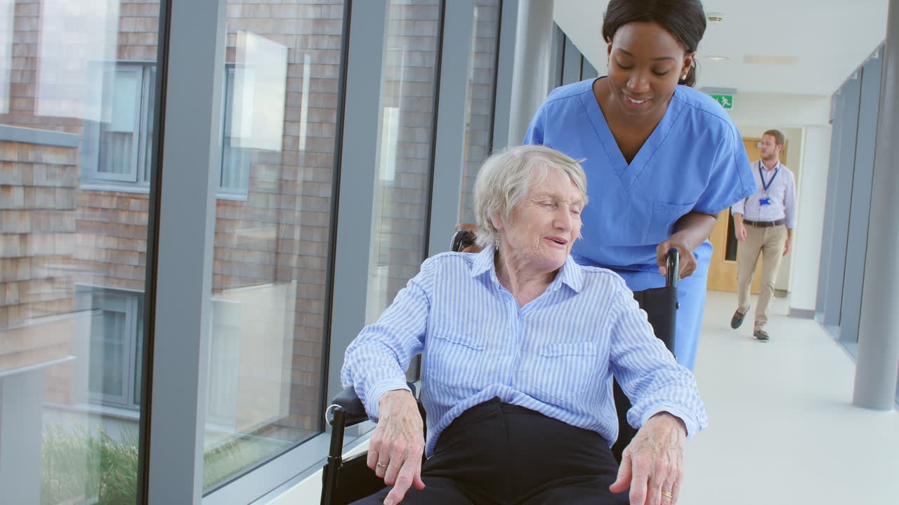 Nurse Pushing Senior Patient In Wheelchair Along Corridor