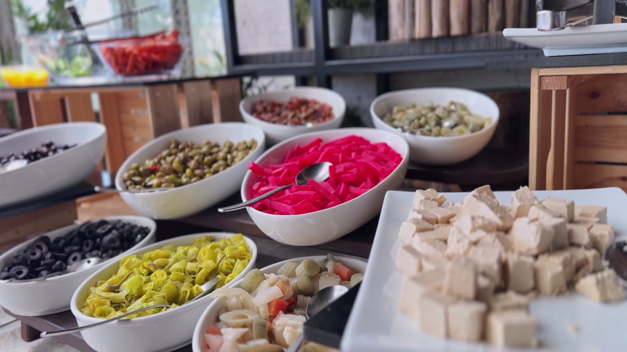 Un colorato buffet mediterraneo con ciotole bianche piene di olive nere e verdi, rape sottaceto rosa, peperoni gialli e cubetti di formaggio su un tavolo di legno scuro all'interno.