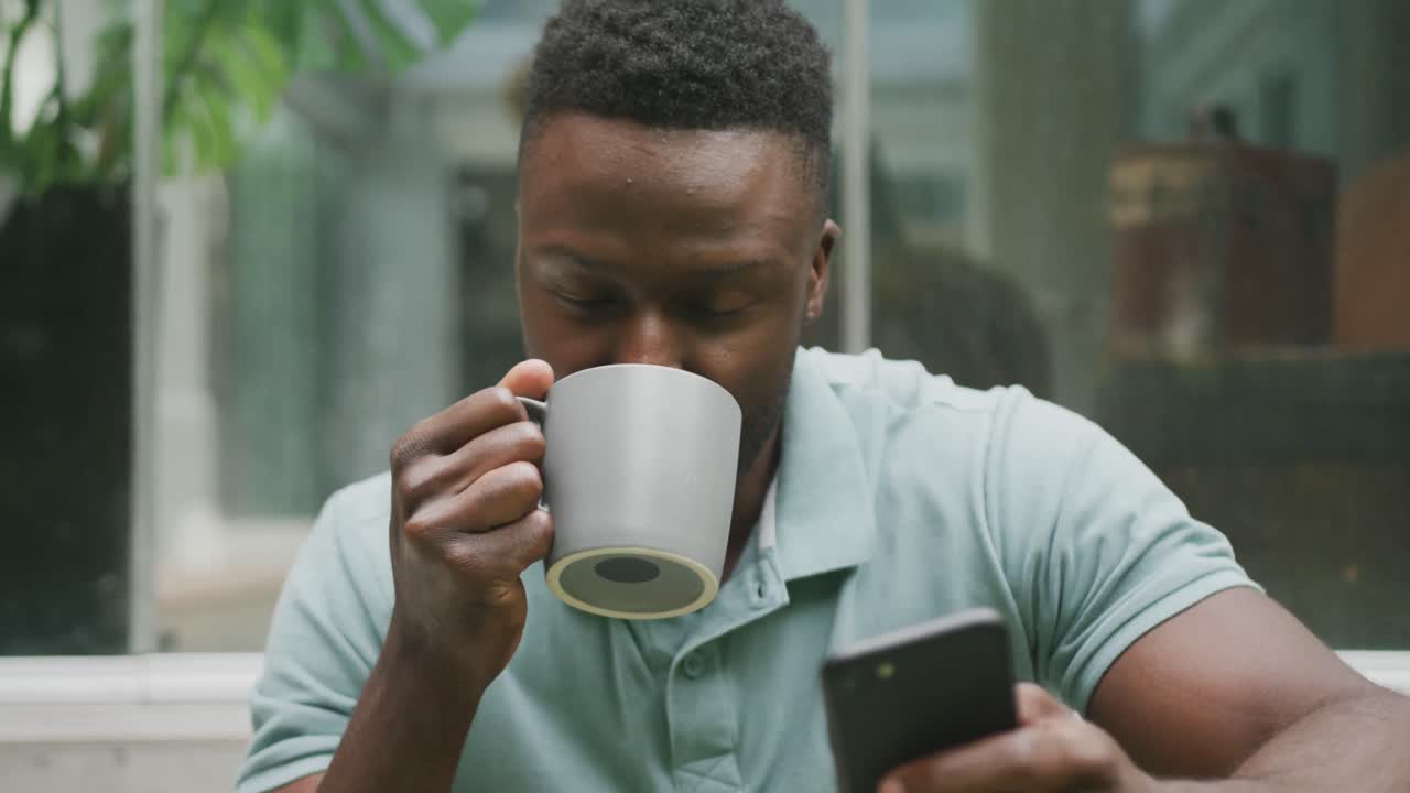 feliz hombre afroamericano sentado con un teléfono inteligente y bebiendo café en el jardín