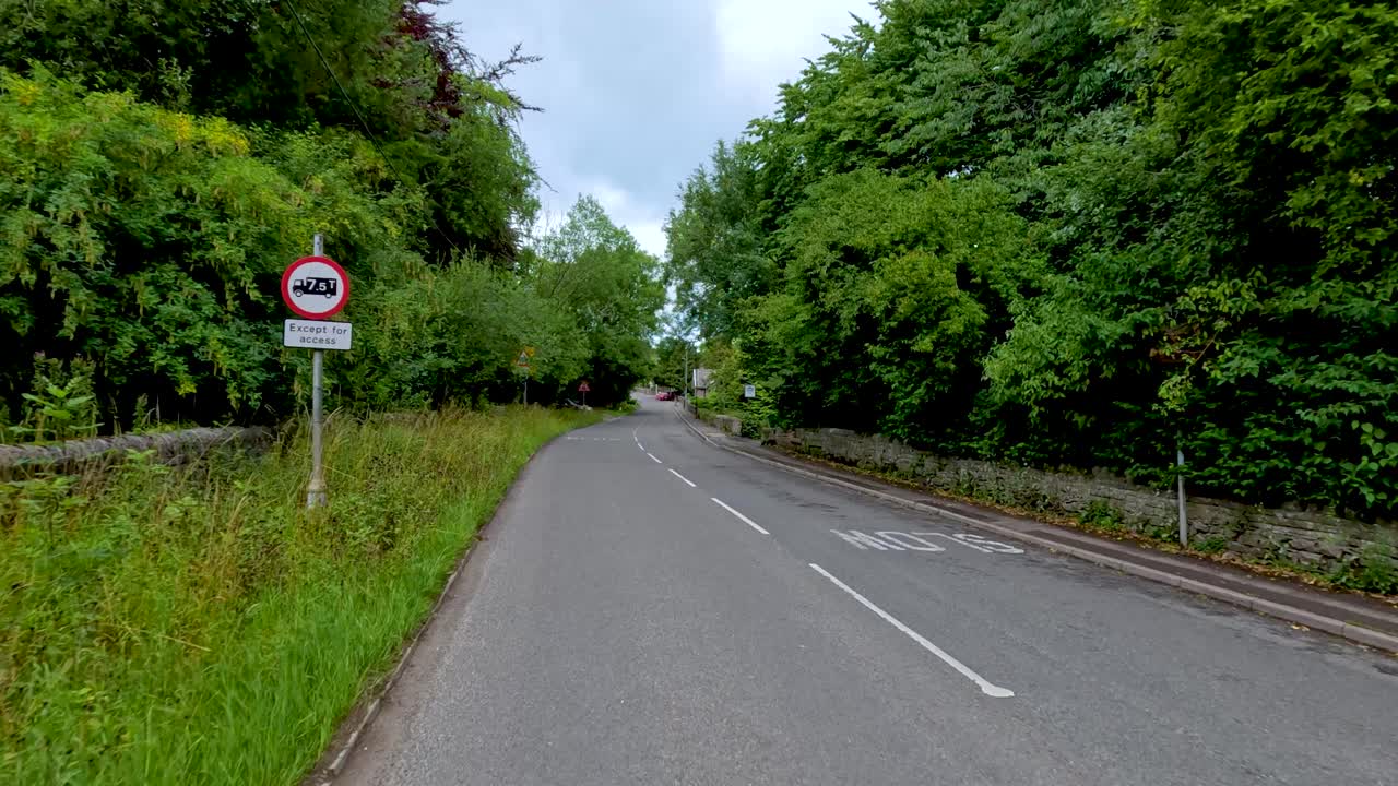 A vehicle travels along a quiet, tree-lined road in the Peak District, England, under overcast daylight, with smooth camera movement and natural surroundings