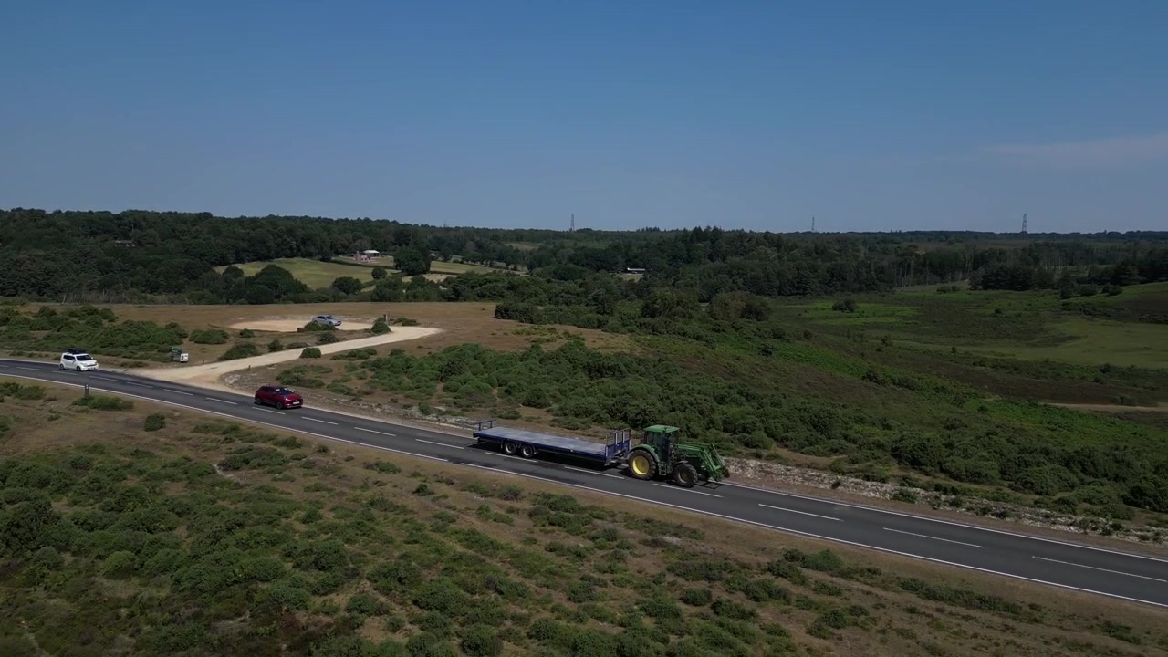 Aerial drone shot of tractor towing trailer along rural road through heathland in New Forest, Hampshire, England with vehicles travelling beneath clear daytime sky