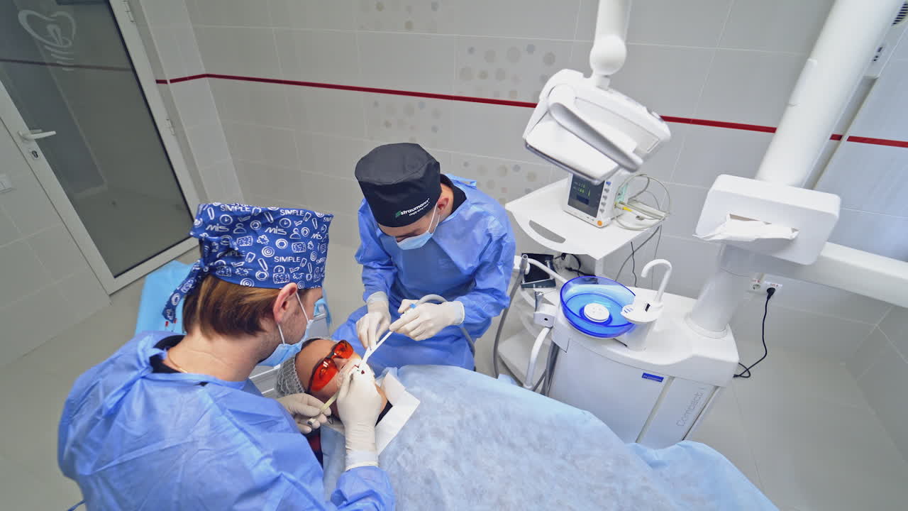 Dentist with assistant treating sick tooth to a patient. Woman patient in protective glasses lying in a dental chair. Top view.