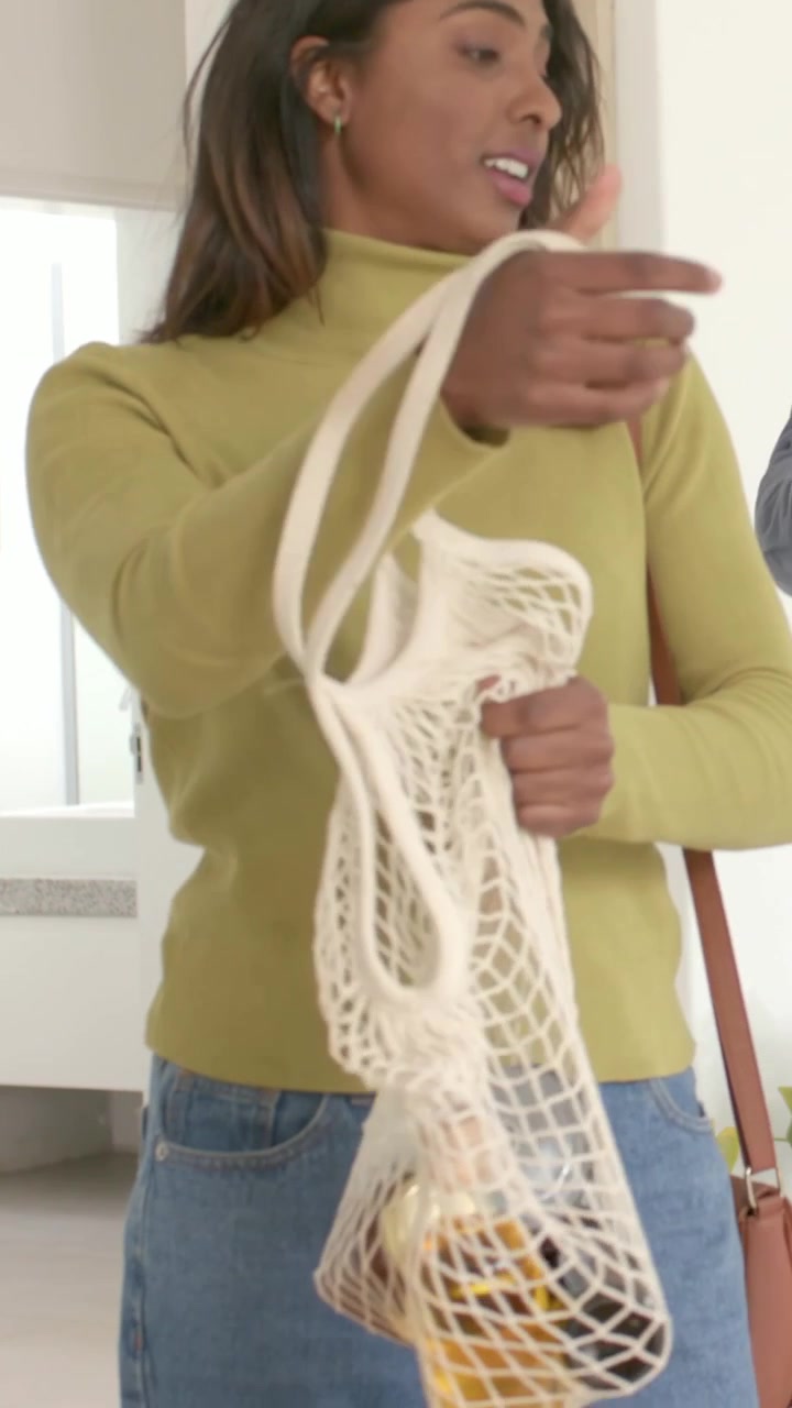 Vídeo vertical: Mulher de blusa verde-oliva ajustando a alça, passando uma sacola de rede com um pote de limões em casa, com espaço para texto.