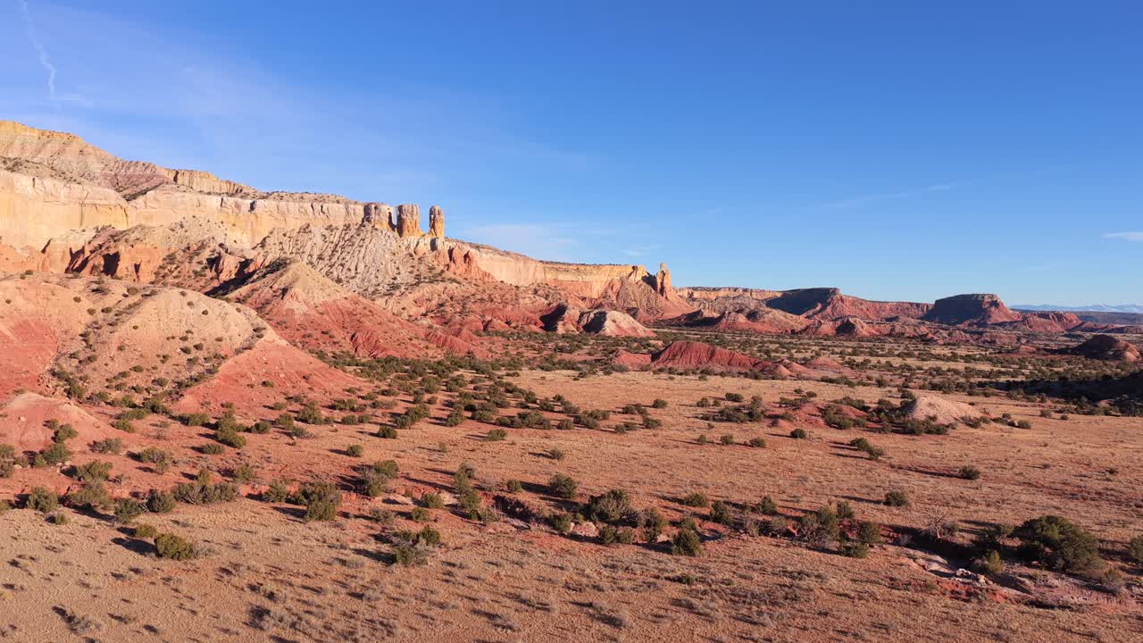Riprese aeree realizzate con un drone che sorvola un vasto e arido territorio desertico, rivelando mesa stratificate di roccia rossa, calanchi erosi, vegetazione scarsa e ampi spazi aperti sotto un cielo azzurro limpido
