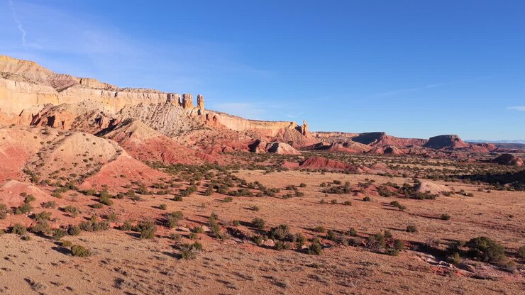 Riprese aeree realizzate con un drone che sorvola un vasto e arido territorio desertico, rivelando mesa stratificate di roccia rossa, calanchi erosi, vegetazione scarsa e ampi spazi aperti sotto un cielo azzurro limpido