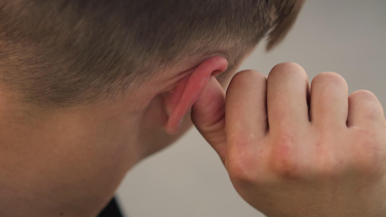 Primer plano de un joven quitándose un auricular detrás de la oreja, con la nuca y los dedos enfocados, ambiente de relajación tras el ejercicio al aire libre, pelo corto informal, movimiento de tirar y alivio, detalle de dispositivo de audio inalámbrico, momento de relajación espontáneo