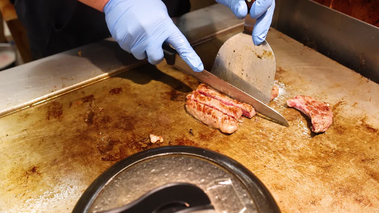 Uno chef che indossa guanti blu usa un coltello e una spatola per affettare manzo wagyu alla griglia.