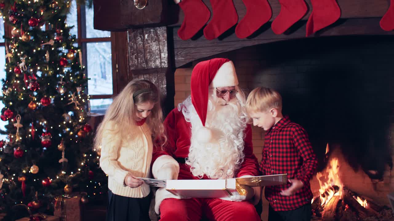 Santa Claus is reading a book to children sitting near a fireplace with Christmas stockings and a Christmas tree
