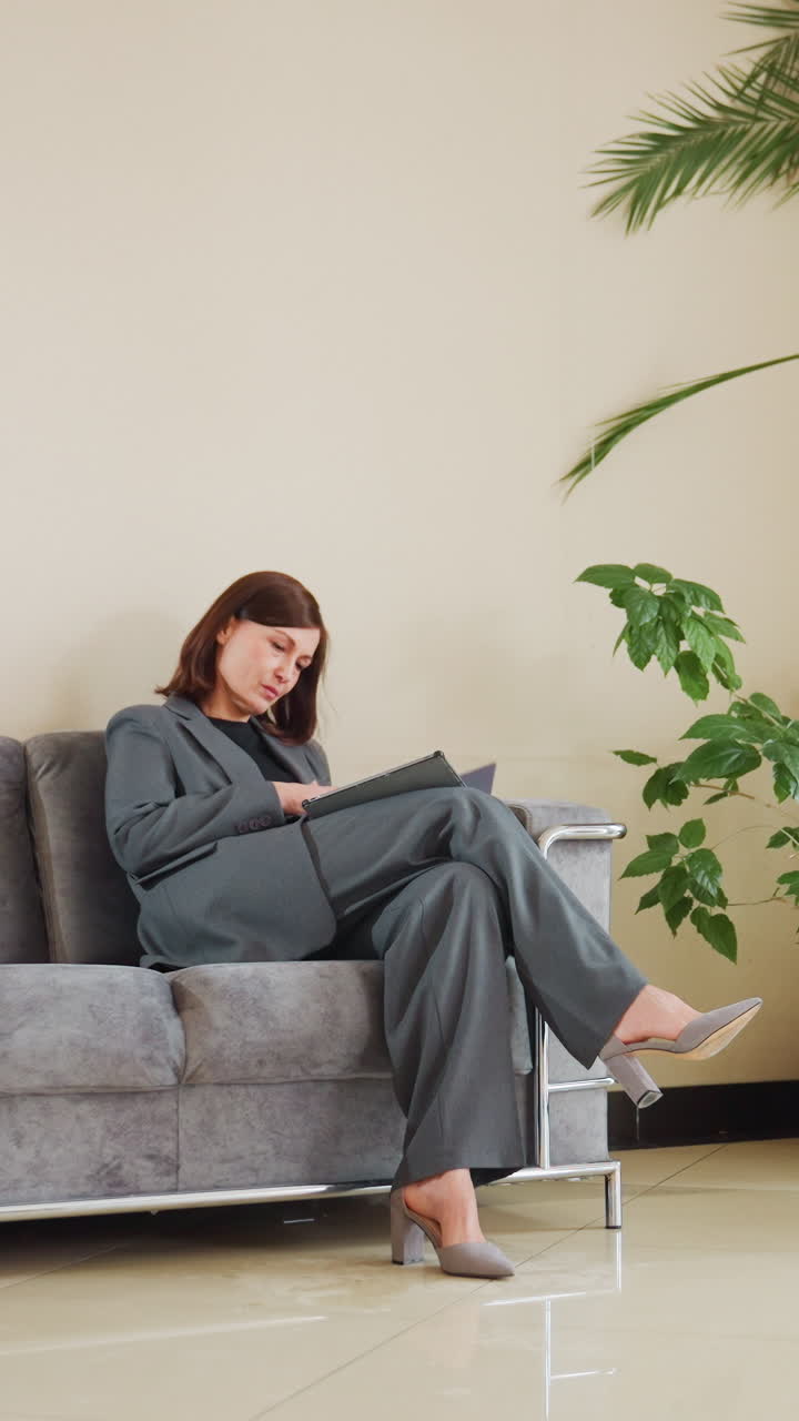 Businesswoman in gray suit stands from couch holding tablet near indoor plants in bright office lobby, appearing ready for meeting or appointment with natural light and modern decor ambiance