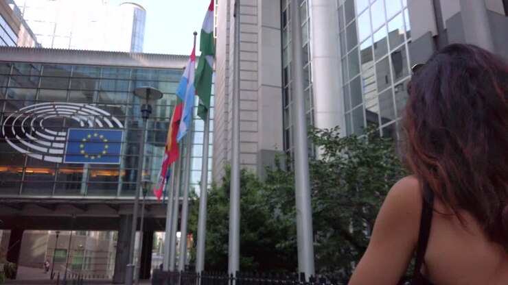 Woman walks toward EU Parliament, flags waving under bright daylight.