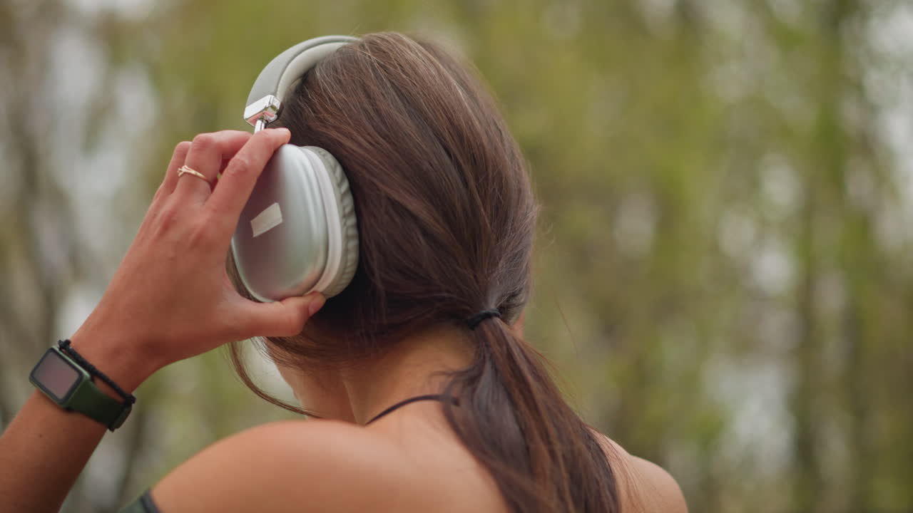 Back view of young girl wearing headphones with a wristwatch on her hand, looking relaxed and connected with nature, capturing a peaceful moment while enjoying music outdoors