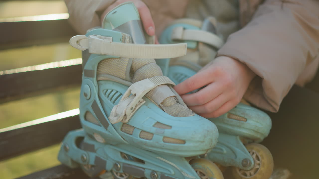 toma de primer plano de una persona con una chaqueta de melocotón sentada en un banco, preparándose para ponerse un par de patines azules claros. se ven las manos de la persona desbloqueando las correas, listas para usar los patines