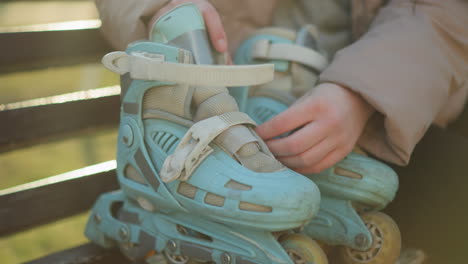 close-up shot of a person in a peach jacket sitting on a bench, preparing to put on a pair of light blue rollerblades. the person's hands are seen unlatching the straps, ready to wear the rollerblades