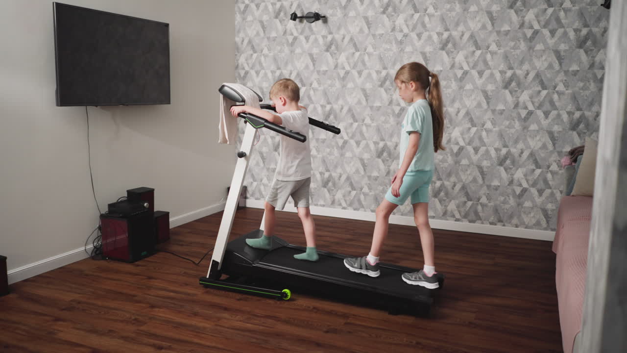 Little boy grips treadmill handle while standing on front of machine as young girl cautiously walks behind, maintaining balance during indoor activity in modern room