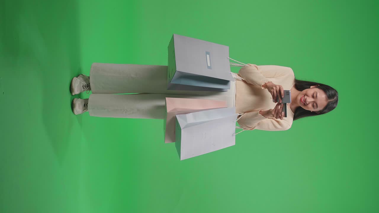 vista completa de una mujer asiática de compras con bolsas de compras y tarjeta de crédito usando el teléfono móvil gastando dinero mientras está de pie frente a la pantalla verde