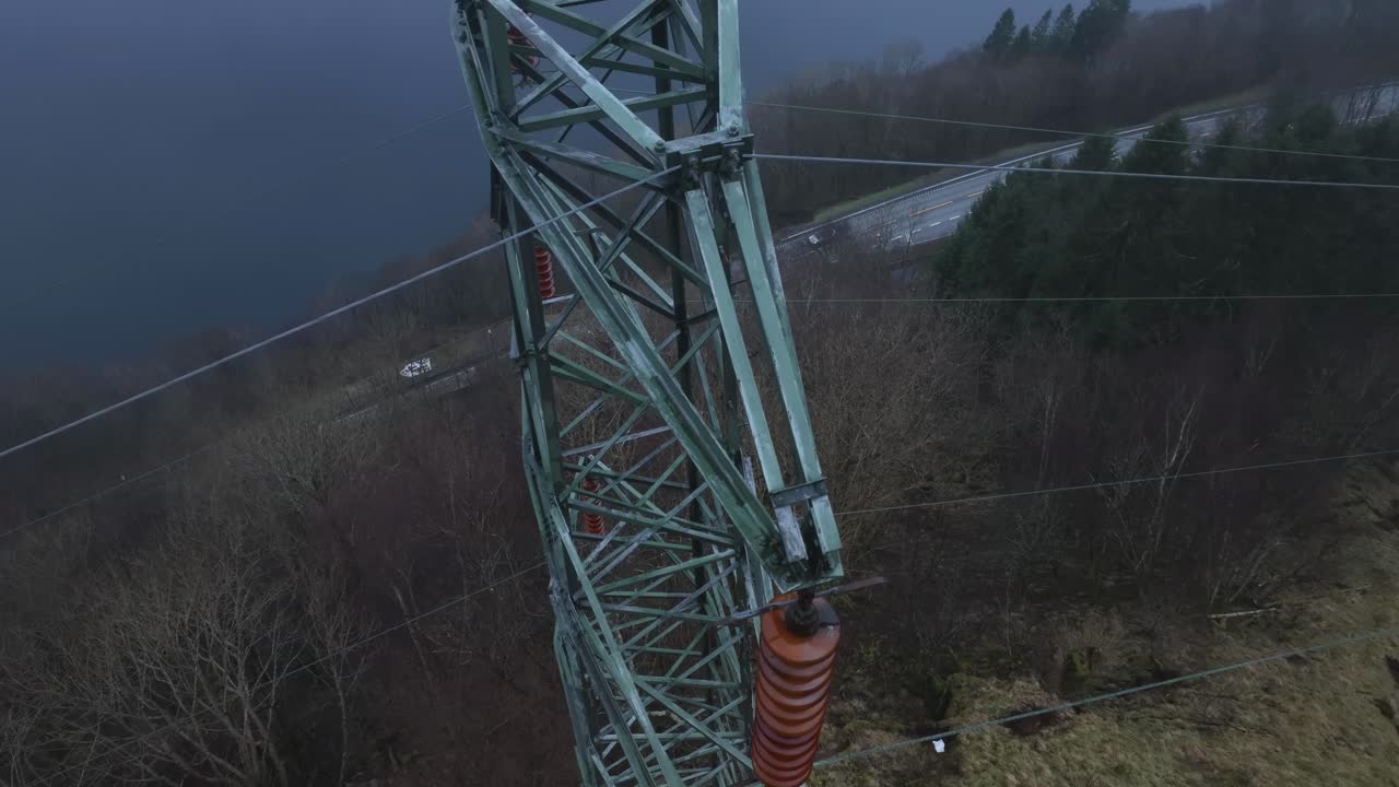 High voltage pylon seen from above, looking down into the forest and a road below. Drone descends while tilting up, revealing Sorfjorden fjord in rainy, misty weather