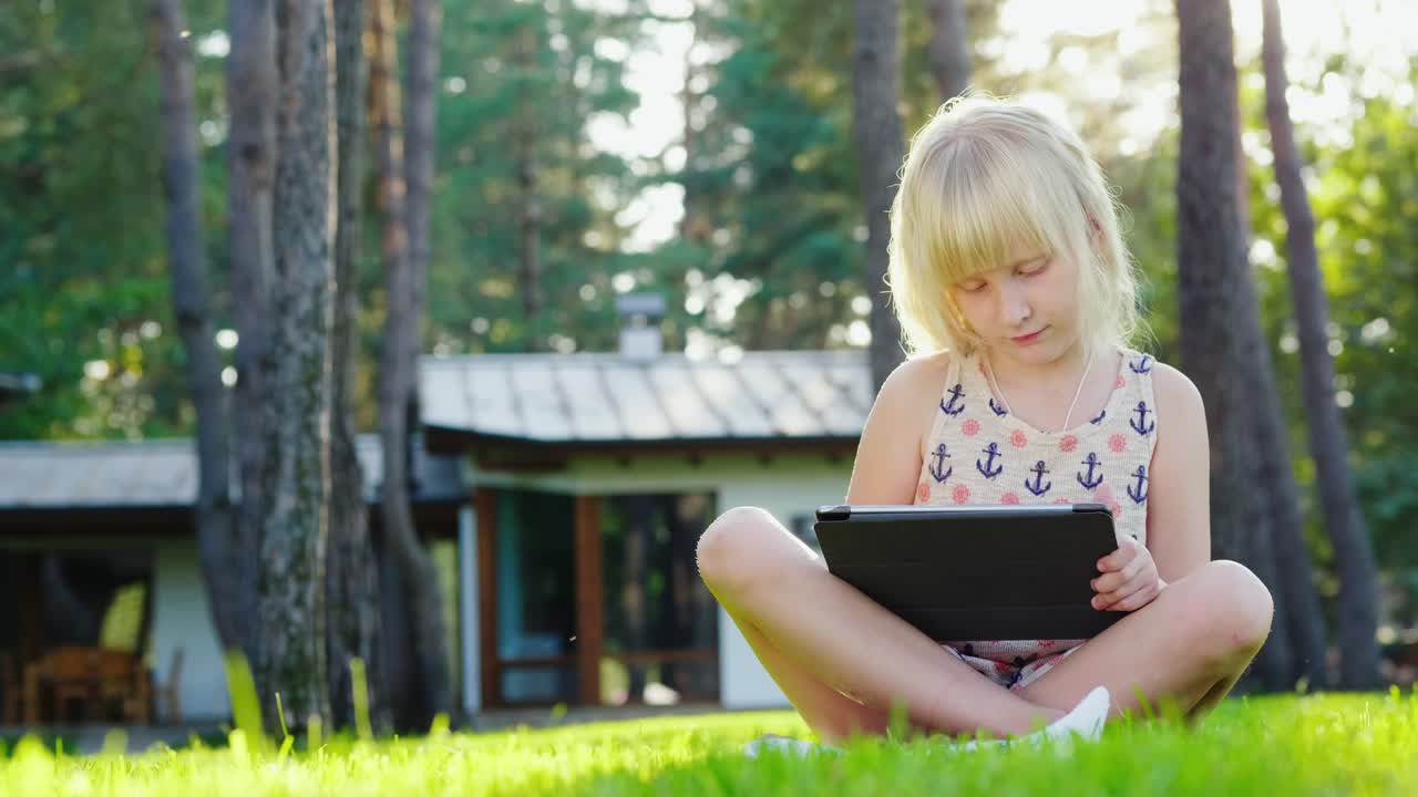 The girl is playing on the tablet. The sun beautifully highlights her blond hair. Sits on the lawn near the house