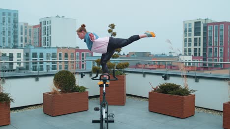 athletic girl performing aerobic riding training exercises on cycling stationary bike on house roof