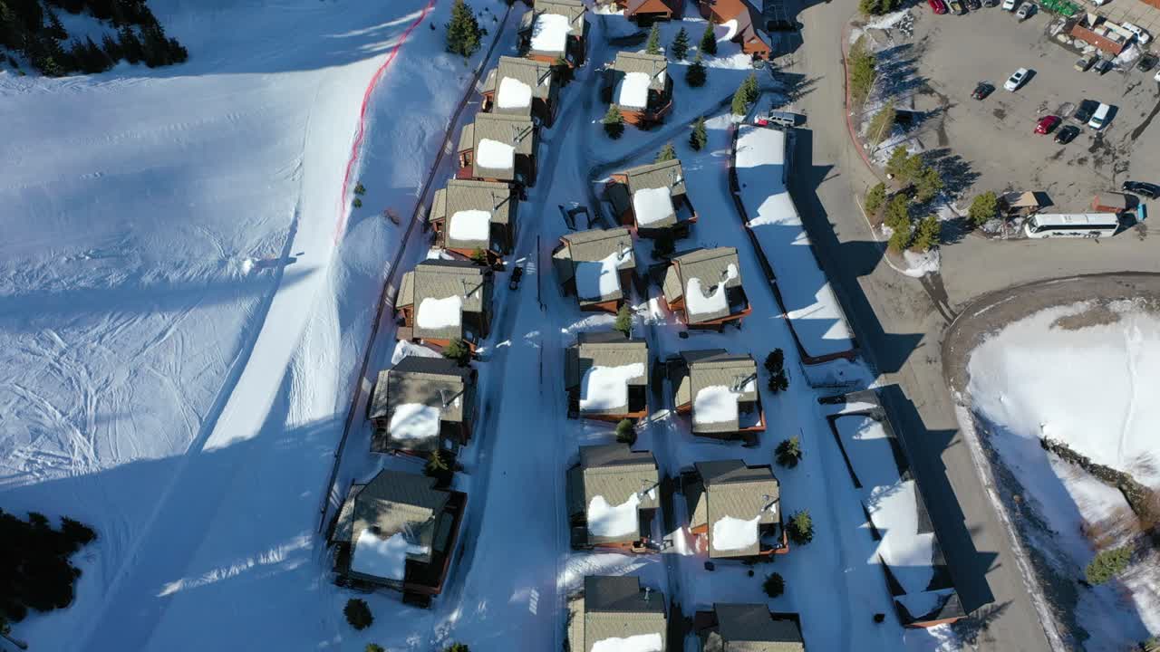 High-altitude drone shot of charming vacation condos in Big Sky Montana, coated in fresh snow, surrounded by evergreens, sloping roads, and vast alpine scenery in a serene winter wonderland
