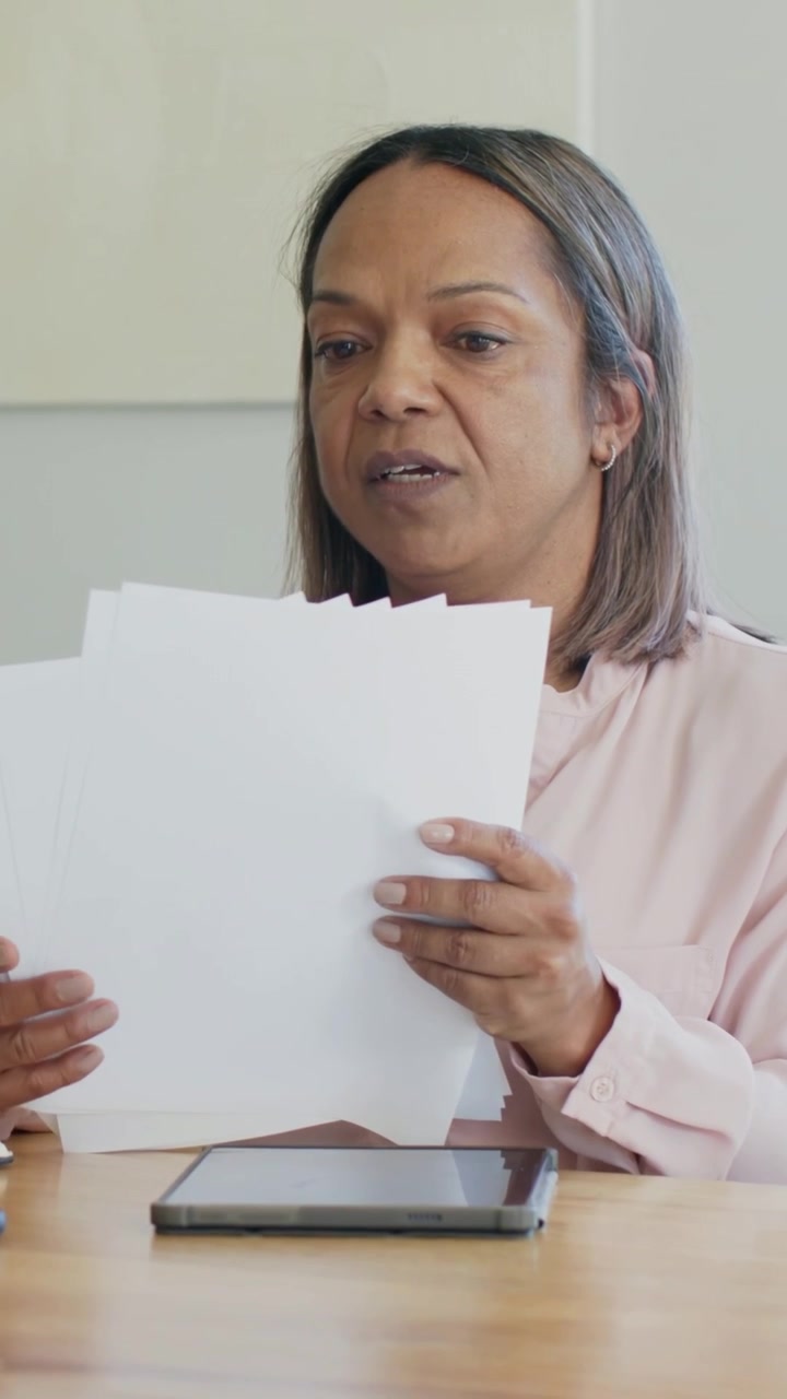 Video verticale: Donna con maglietta rosa che sventaglia una risma dopo averla raccolta, leggendo in ufficio con un tablet per lavoro.