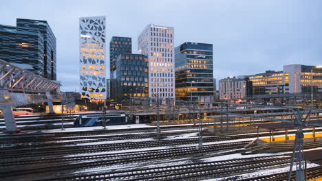 Train-Travelling-Under-Acrobat-Bridge-With-Illuminated-Buildings-On-Barcode-Project-At-Oslo,-Norway