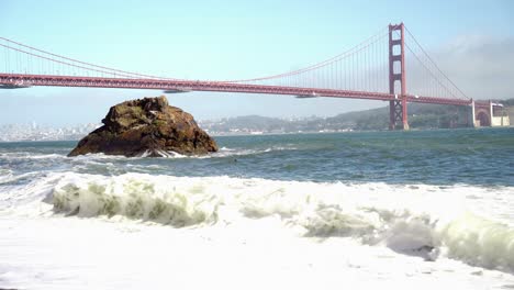 view of the golden gate bridge and waves crashing on the shore and a big rock