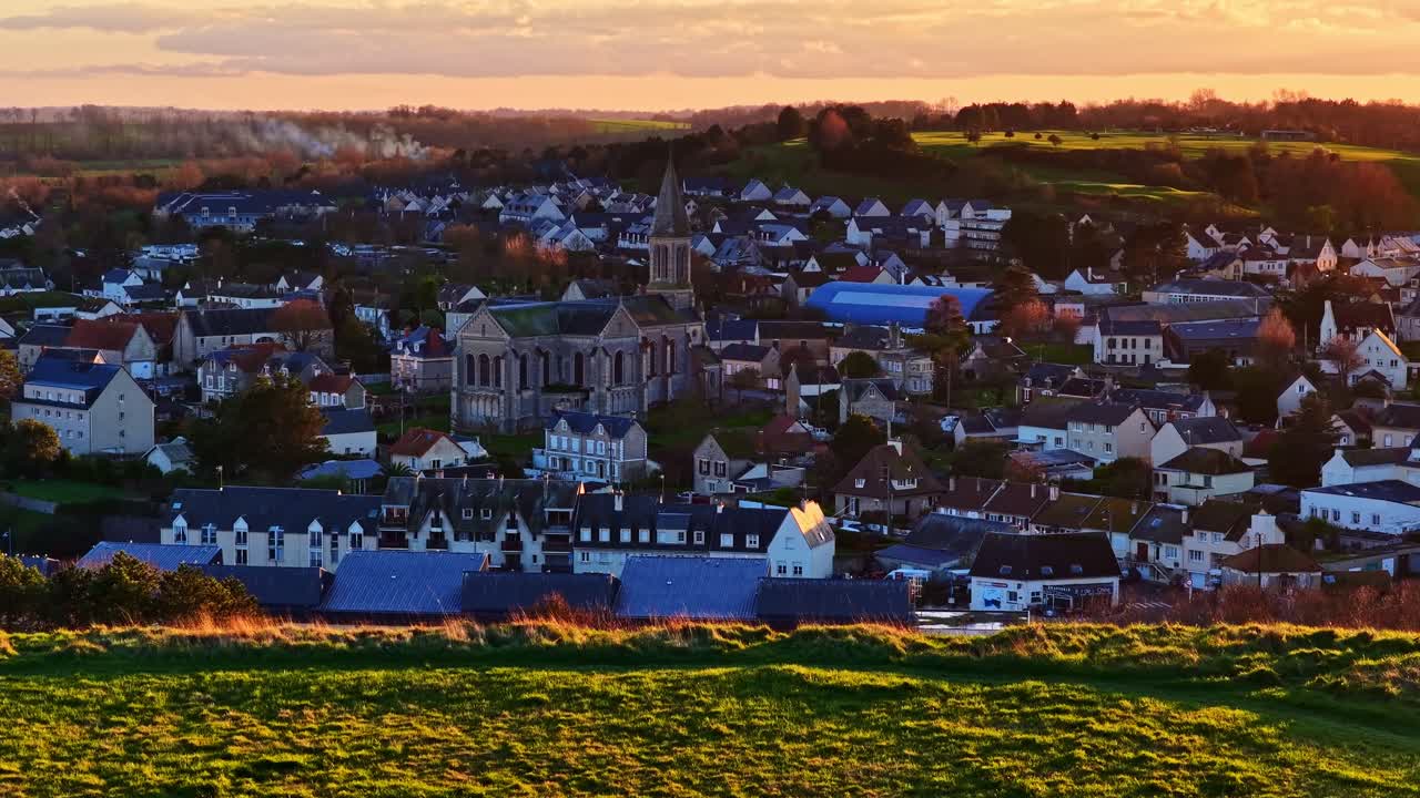 I tetti e il campanile di una città della Normandia risplendono caldamente al tramonto sulla campagna aperta. Francia, Port en Bessin.