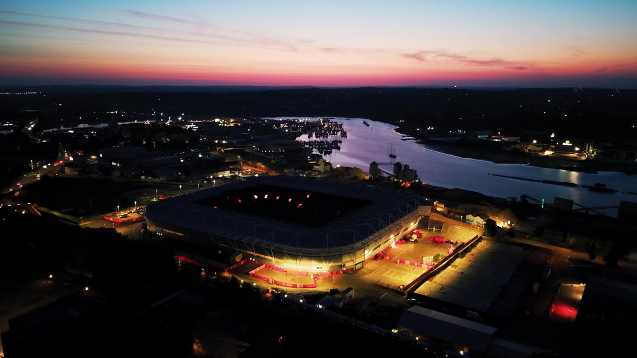 Høj droneudsigt over det oplyste St Marys Stadium, byens lys og floden Itchen under en levende lyserød horisont før daggry.