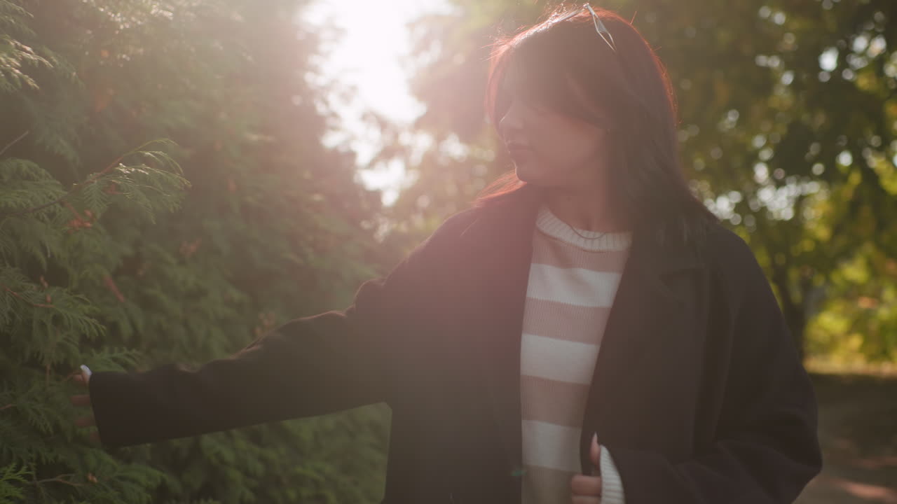 Dim view of autumn visitor touching through with her hands green leaf, soft sun flare over hedge, striped sweater under dark coat, calm walk along park pathway, serene outdoor travel vibe