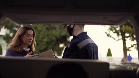 hermosa mujer y hombre de entrega en una máscara protectora y guantes abriendo el maletero y tomando el paquete de caja de cartón. mensajero