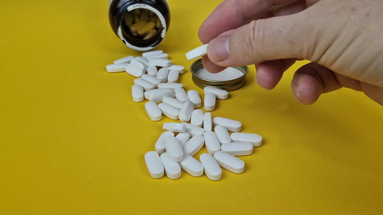 Macro 4K shot of a hand placing a white, oblong pill next to an open, brown black prescription or supplement bottle and a scattered pile of pills on a vibrant yellow background