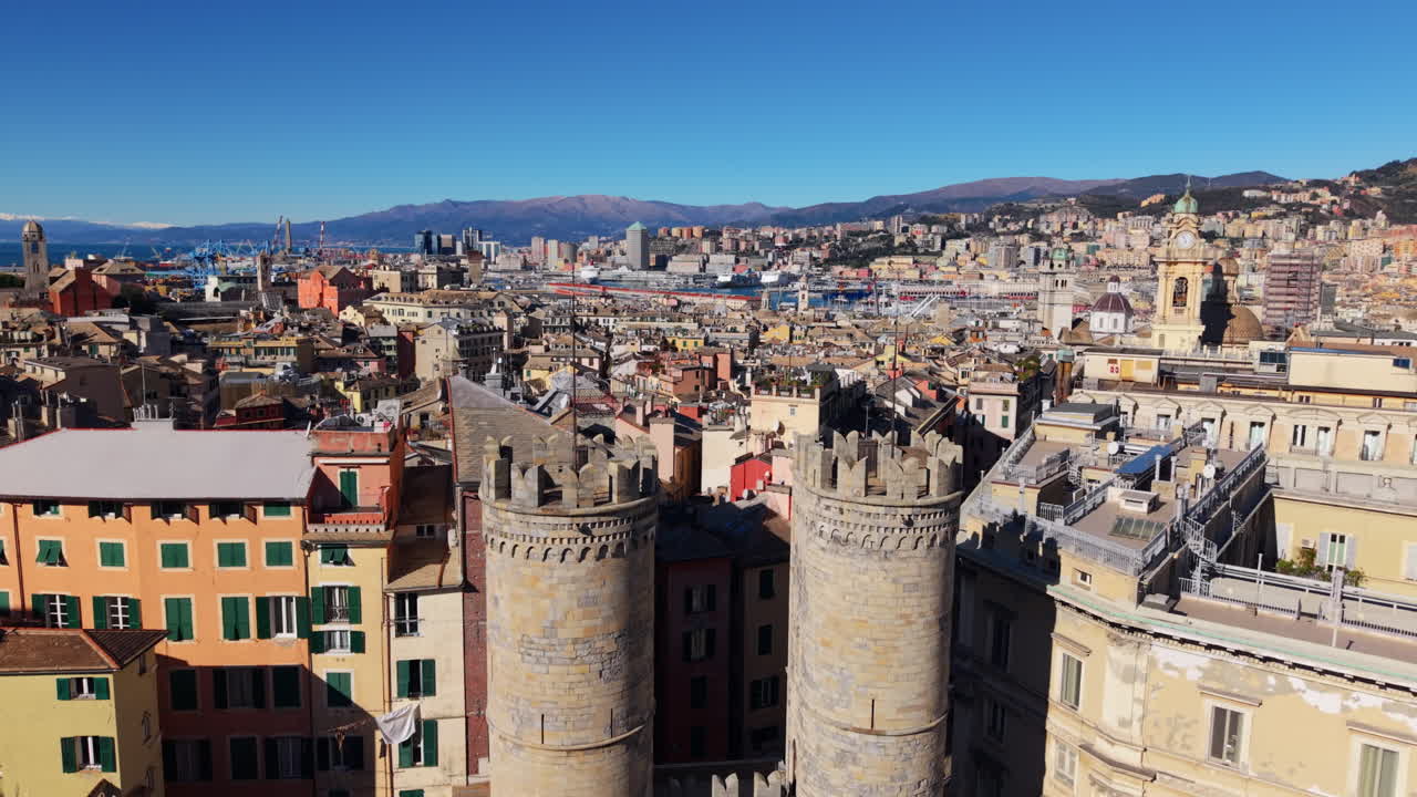 Un drone s'élève devant les tours de la Porta Soprana à Gênes, en Italie, révélant le centre historique dense, des bâtiments colorés, la zone portuaire et les montagnes ligures sous un ciel dégagé