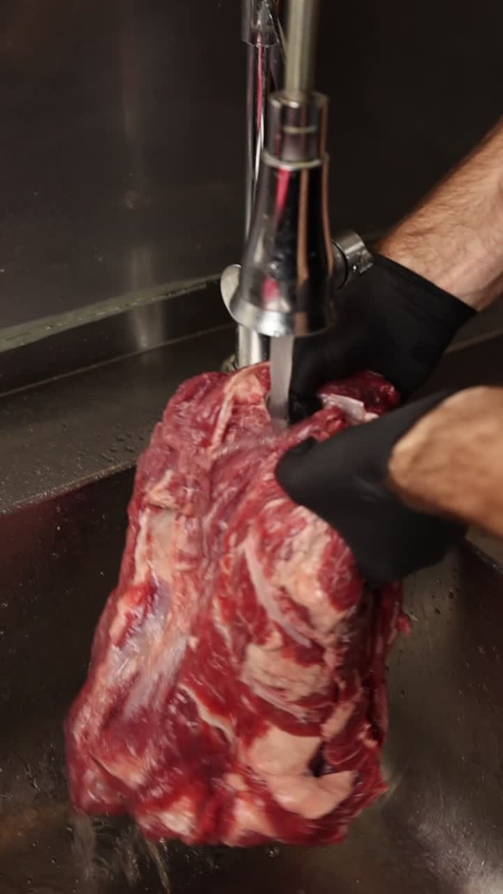 Mani con guanti che lavano un grosso taglio di manzo crudo nel lavello della cucina