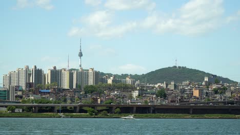 Yacht,-Die-Auf-Dem-Fluss-Han-Treibt,-Autoverkehr-Auf-Der-Brücke-Namsan-Tower-Und-Villen-Und-Wohnungen-Im-Yongsan-viertel-Im-Hintergrund-An-Einem-Sonnigen-Bewölkten-Tag,-Statisch