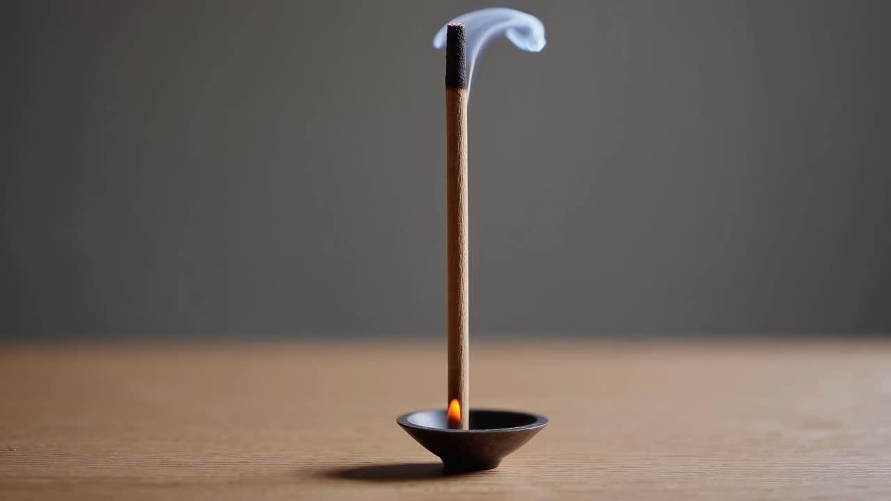 A close-up video of a burning incense stick in a small holder on a wooden surface