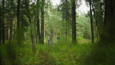 little girl lets go of mother hand to collect object from brother while walking through dense forest, surrounded by tall pine trees, green foliage, and soft light filtering through woodland scenery