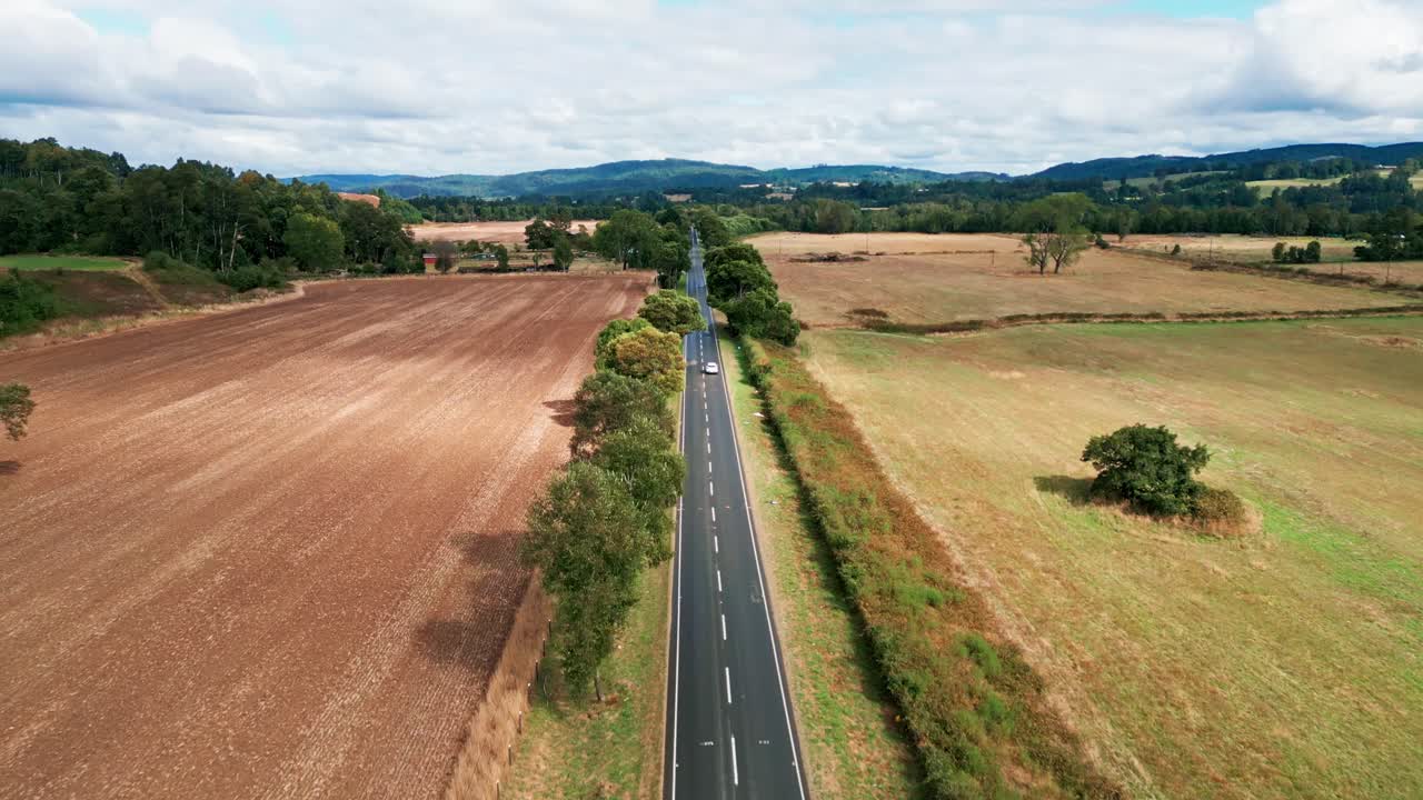 Drohnenflug verfolgt Auto auf gerader Straße durch landwirtschaftliche Felder.
