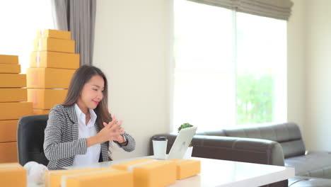 Asian-businesswoman-has-done-all-orders-for-shipping-and-looking-at-work-on-laptop-computer-with-satisfaction,-stretching-arms-in-the-air-with-closed-eyes-and-happiness,-surrounded-with-yellow-boxes