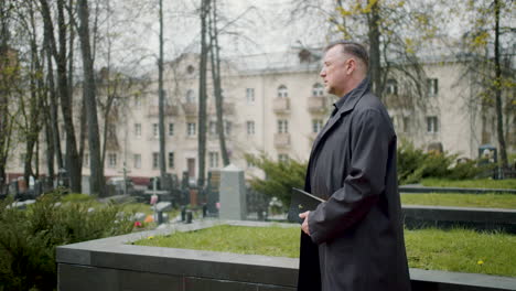 hombre con impermeable negro sosteniendo una biblia y mirando el horizonte en un cementerio