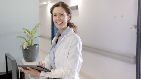 portrait of happy caucasian female doctor with tablet at hospital reception, copy space, slow motion