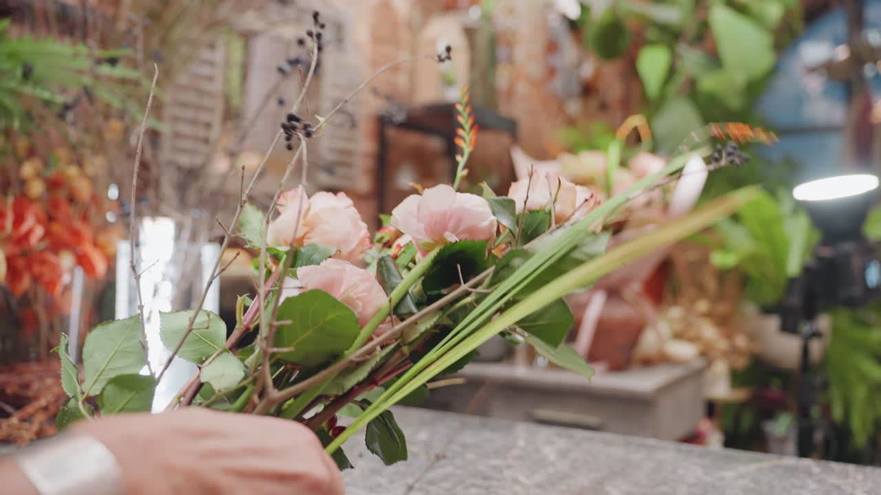 Mani che assemblano un bouquet stagionale, fiorista caucasica, steli e verde disposti sul banco da lavoro, rifinitura e legatura ritmiche, sfondo vivace del mercato, lavoro artigianale da studio e composizione floreale fresca