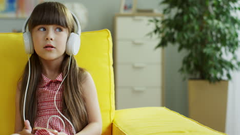 Niña-Linda-Sentada-En-Un-Sofá-Amarillo-Escuchando-Música-Con-Auriculares,-Cantando-Y-Bailando-Sentada-En-Un-Sofá-En-La-Sala-De-Estar-En-Casa