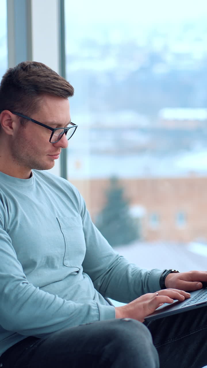 Busy focused man in glasses looks attentively at the screen of his laptop. Freelancer distracts from work for a moment glancing into window. Vertical video
