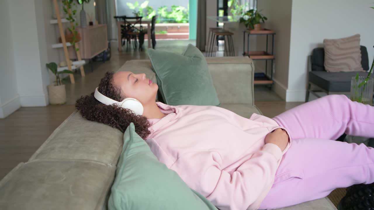 African American woman in pink hoodie lounging on sofa at home tapping phone fixing headset resting