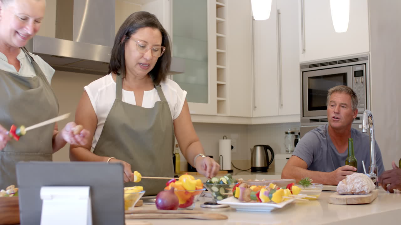 In kitchen, senior diverse women preparing vegetable skewers together, focusing on cooking, at home