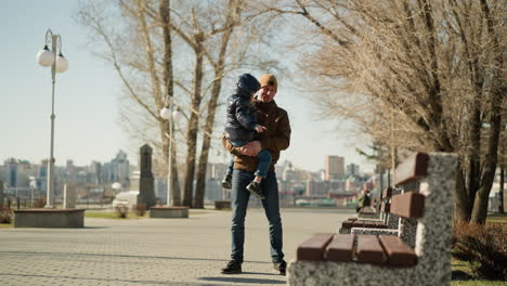 father carrying his son while standing near a bench on a paved pathway, with an urban cityscape in the background, the child is dressed in a puffy jacket