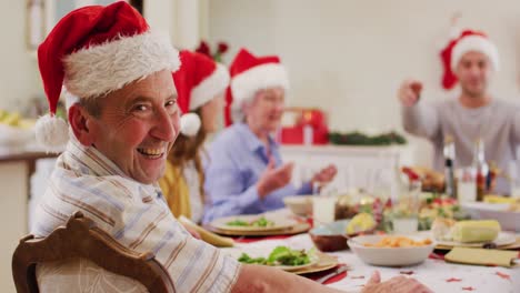 Portrait-of-caucasian-senior-man-in-santa-hat-smiling-while-sitting-on-dining-table-and-enjoying-lun